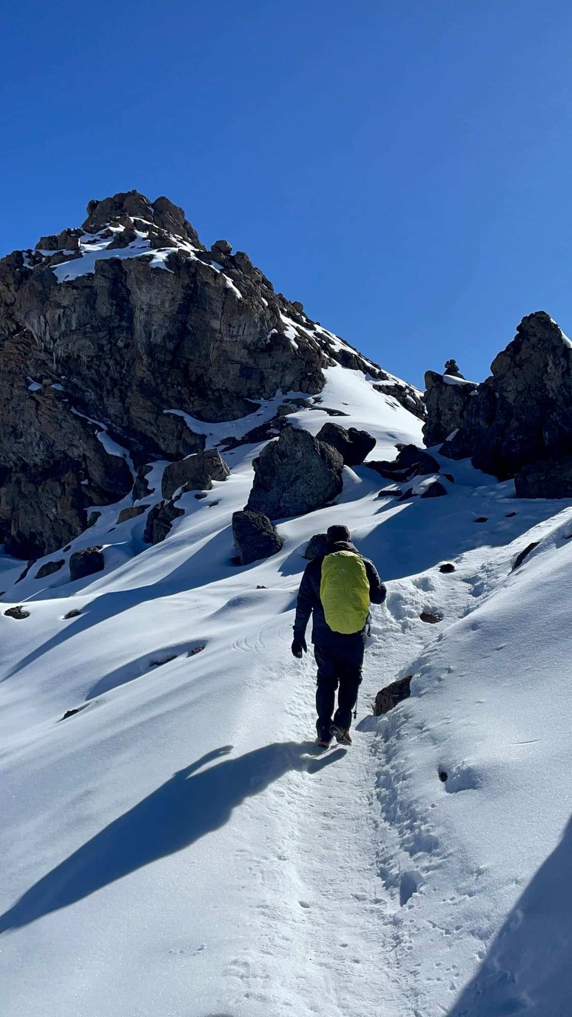 A climber with a yellow pack ascending a snowed-in trail toward rocky pinnacles
