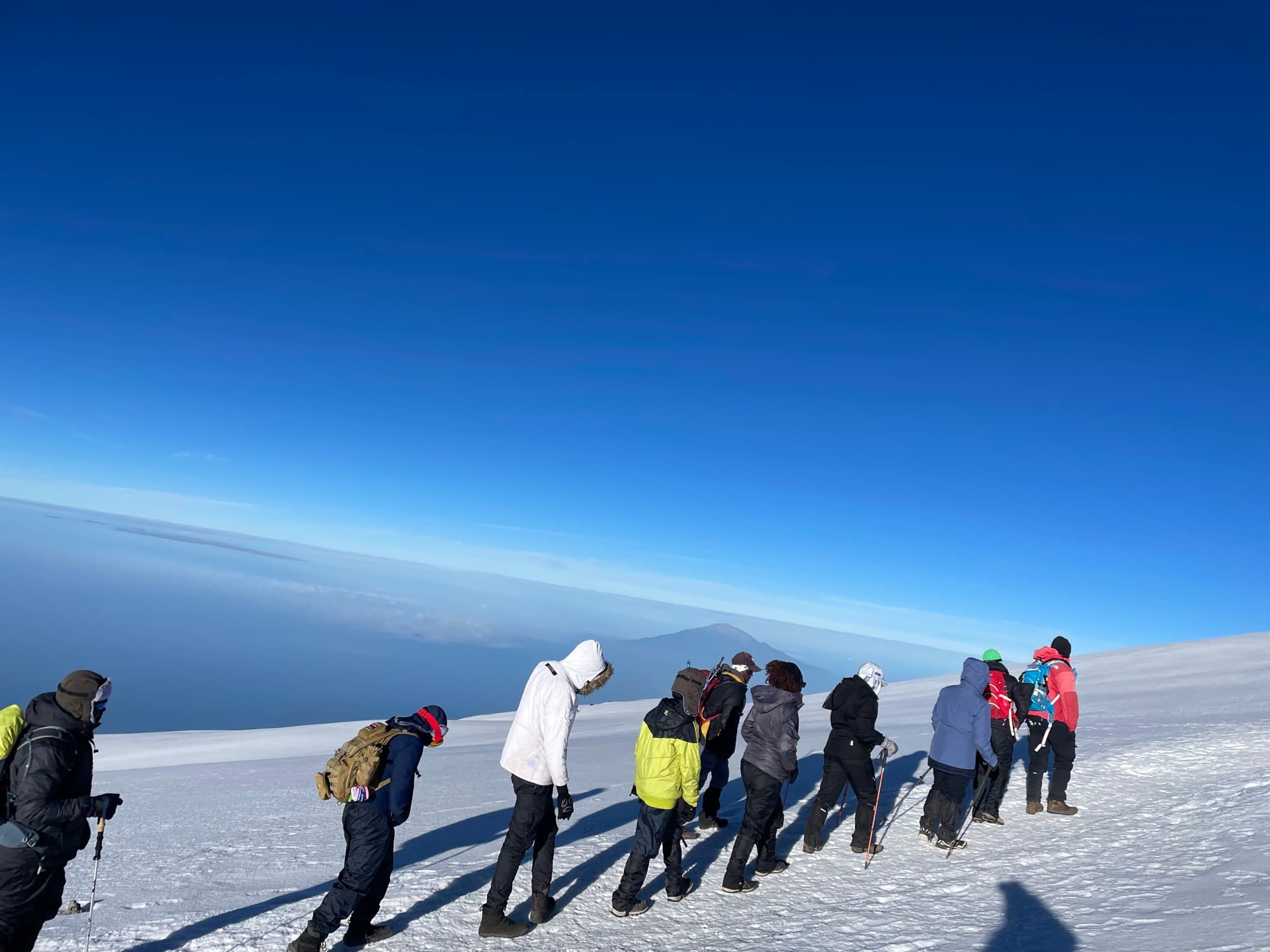 A rope of trekkers crossing the crater-rim snowfield at dawn with Meru beyond